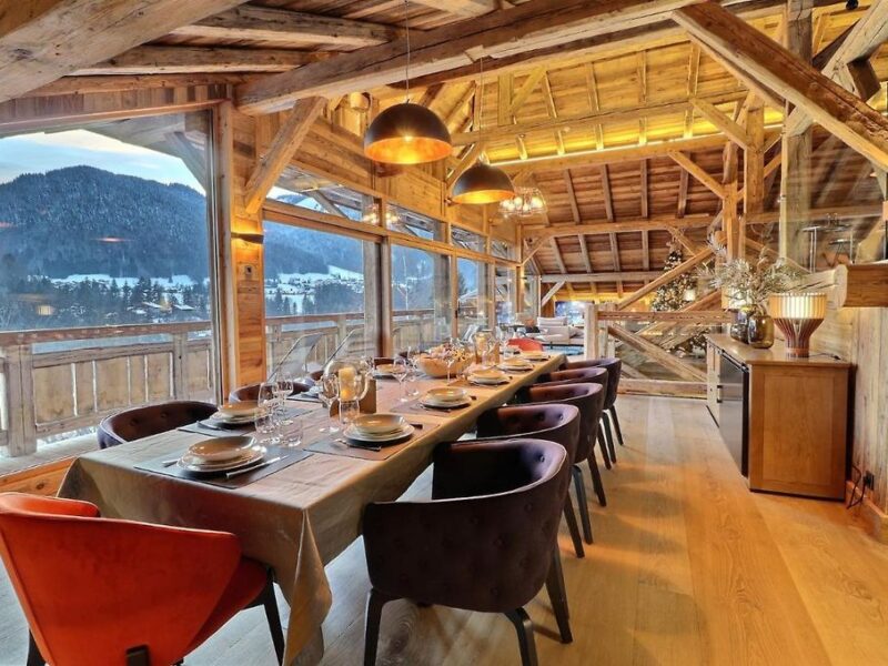 Salle à manger d’un chalet à Morzine avec éclairage chaleureux, poutres apparentes et vue sur les montagnes enneigées